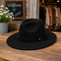 A black felt wide-brim fedora hat with a matching black band and small gold button detail, displayed on a rustic wooden table in a boutique setting.
