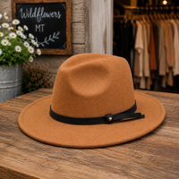 A camel brown felt fedora hat with a black ribbon band and a small gold button, displayed on a rustic wooden table in a shop setting.