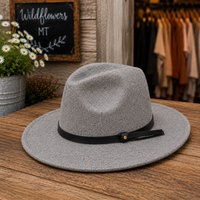 A gray felt fedora hat with a black band and small gold button, displayed on a wooden table in a rustic shop setting with flowers and clothing racks in the background.