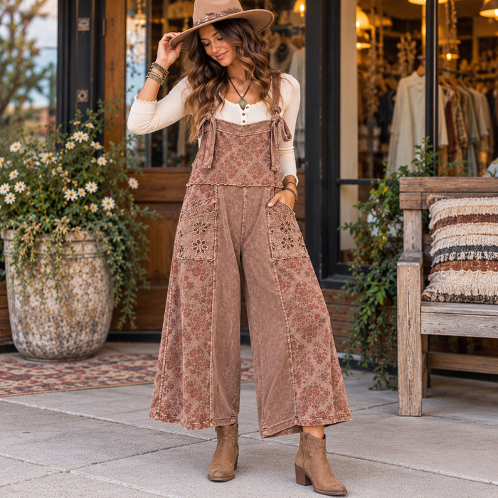 A woman stands on a storefront sidewalk wearing a brown floral wide-leg overall jumpsuit over a cream long-sleeve top, with a tan hat and brown ankle boots. The shop entrance, potted flowers, and bench are visible in the background.