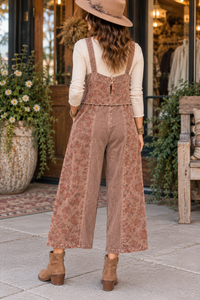 A woman stands on a sidewalk in front of a rustic storefront, viewed from behind. She is wearing dusty rose-brown floral wide-leg corduroy overalls over a cream long-sleeve top, a tan wide-brim hat, and tan ankle boots.