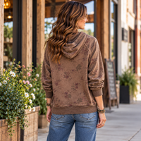 A woman viewed from behind wearing a brown hooded sweatshirt with a faded floral pattern, paired with blue jeans, standing on a sidewalk in front of a rustic storefront.