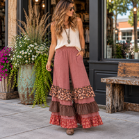 A woman stands on a sidewalk in front of a storefront, wearing wide-leg pleated pants in dusty rose with layered floral and checkered ruffle hems, paired with a cream sleeveless top and brown boots.