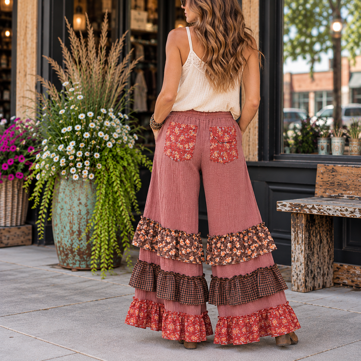 A woman stands on a storefront sidewalk wearing loose, wide-leg dusty rose pants with floral and plaid ruffle hems and matching floral back pockets. The photo is taken from behind, with a shop window, potted plants, and bench visible in the background.