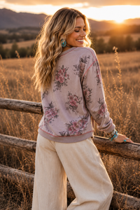 A woman stands outdoors in a sunlit field near a wooden fence, wearing a long-sleeve floral sweatshirt and cream wide-leg pants. The background shows dry grass, distant hills, and a warm sunset glow.