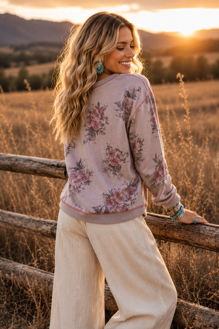 A woman stands outdoors in a sunlit field near a wooden fence, wearing a long-sleeve floral sweatshirt and cream wide-leg pants. The background shows dry grass, distant hills, and a warm sunset glow.