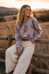 A woman sitting on a rustic wooden fence in a golden field at sunset, wearing a dusty mauve floral long-sleeve sweatshirt, cream wide-leg pants, and brown boots.