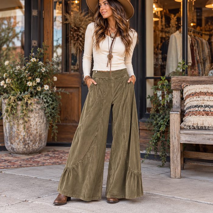 A woman stands on a sidewalk in front of a boutique doorway wearing wide-leg olive corduroy pants, a cream long-sleeve top, a tan hat, and brown boots. The setting includes potted plants, a wooden bench with a woven cushion, and store fixtures.