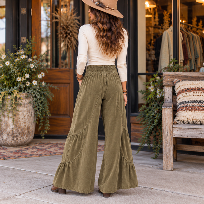 A woman stands with her back to the camera wearing wide-leg olive green pants, a cream long-sleeve top, and a tan hat in front of a rustic storefront with potted plants and a wooden bench.