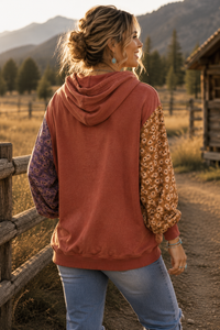 A woman standing outdoors in a rustic mountain setting, wearing a rust-colored hooded sweatshirt with patterned purple and tan sleeves, paired with blue jeans.