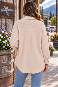 A woman viewed from behind wearing a light beige ribbed button-up shirt and blue jeans, standing outdoors on a sidewalk in front of a rustic wooden building with flowers and mountains in the background.