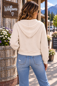 A woman viewed from behind wearing a cream waffle-knit cropped hoodie and light blue jeans, standing outdoors in front of a rustic wooden barrel planter and a sign that reads 'Wildflowers'.