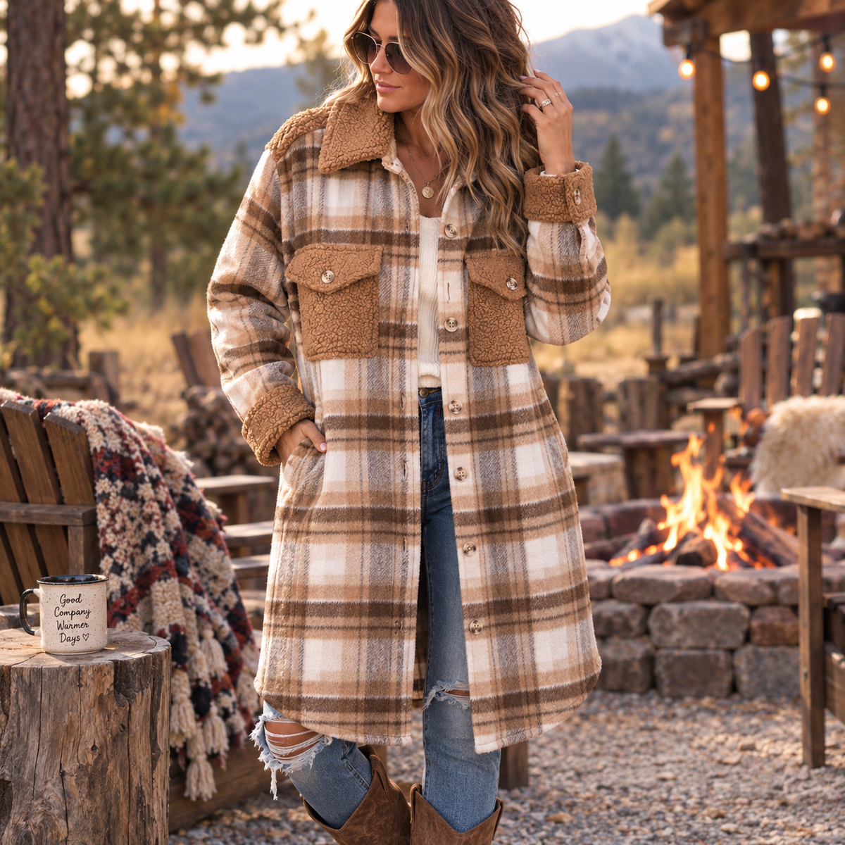 A woman wearing a long beige and brown plaid coat with a brown sherpa collar stands outdoors near a campfire at dusk. The coat has large front flap pockets and button-front closure, paired with blue jeans and brown boots in a rustic lodge-like setting.