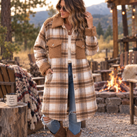 A woman wearing a long beige and brown plaid coat with a brown sherpa collar stands outdoors near a campfire at dusk. The coat has large front flap pockets and button-front closure, paired with blue jeans and brown boots in a rustic lodge-like setting.
