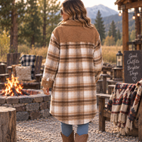 A woman stands with her back to the camera wearing a mid-length plaid fleece jacket with a brown sherpa collar, in an outdoor rustic campfire setting.