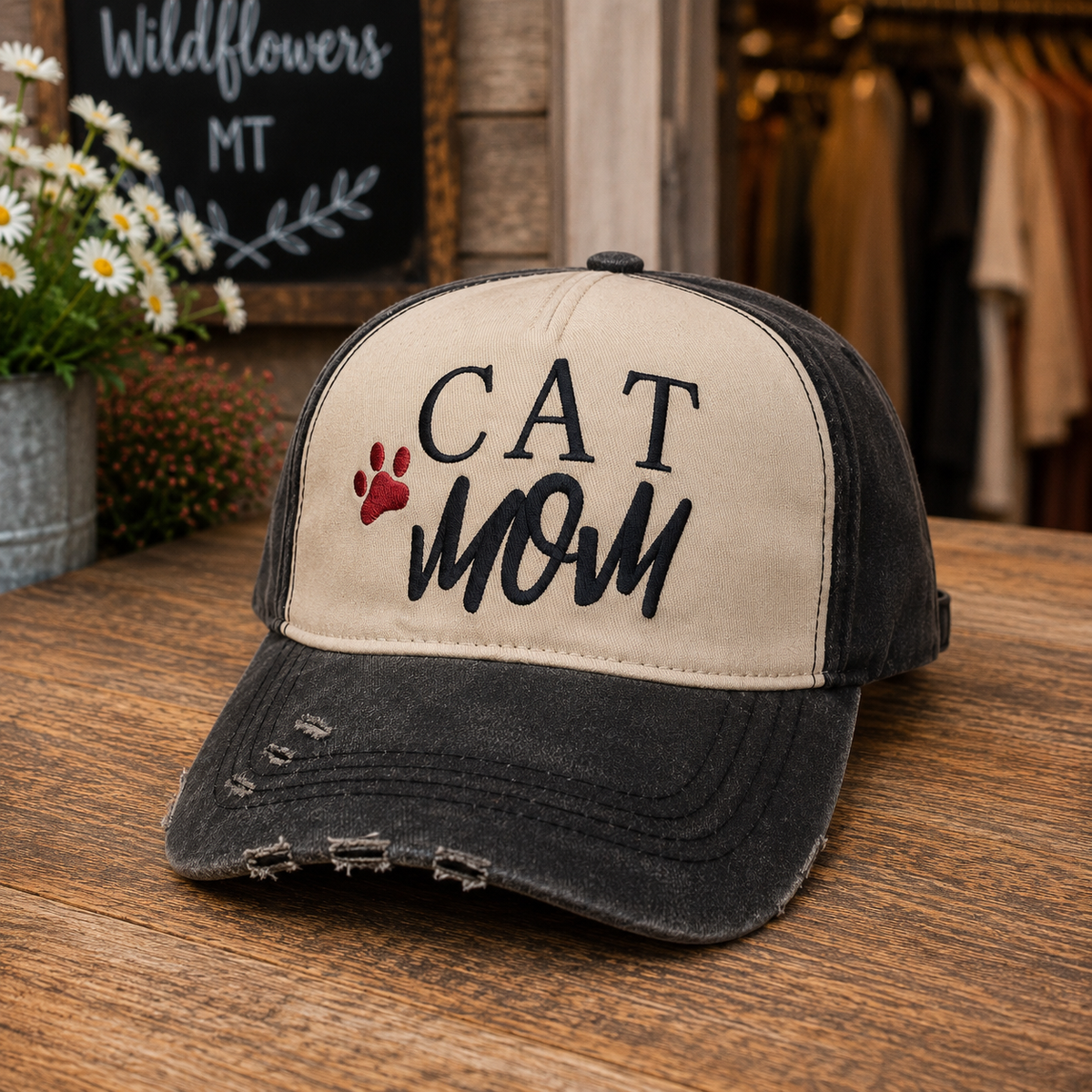 A distressed trucker-style baseball cap with a beige front panel and dark charcoal gray brim, featuring the embroidered text 'CAT MOM' and a small red paw print. The cap is displayed on a rustic wooden table with a blurred boutique background.