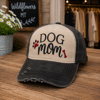 A distressed baseball cap with a cream front panel and a dark gray washed brim, featuring the words 'DOG mom' in black script with red paw print and bone accents. The cap is displayed on a wooden table in a store setting.
