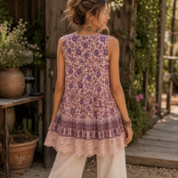 A woman wearing a sleeveless bohemian-style tunic top with a purple and dusty pink floral pattern, lace hem, and crochet yoke, photographed from behind in an outdoor rustic garden setting.