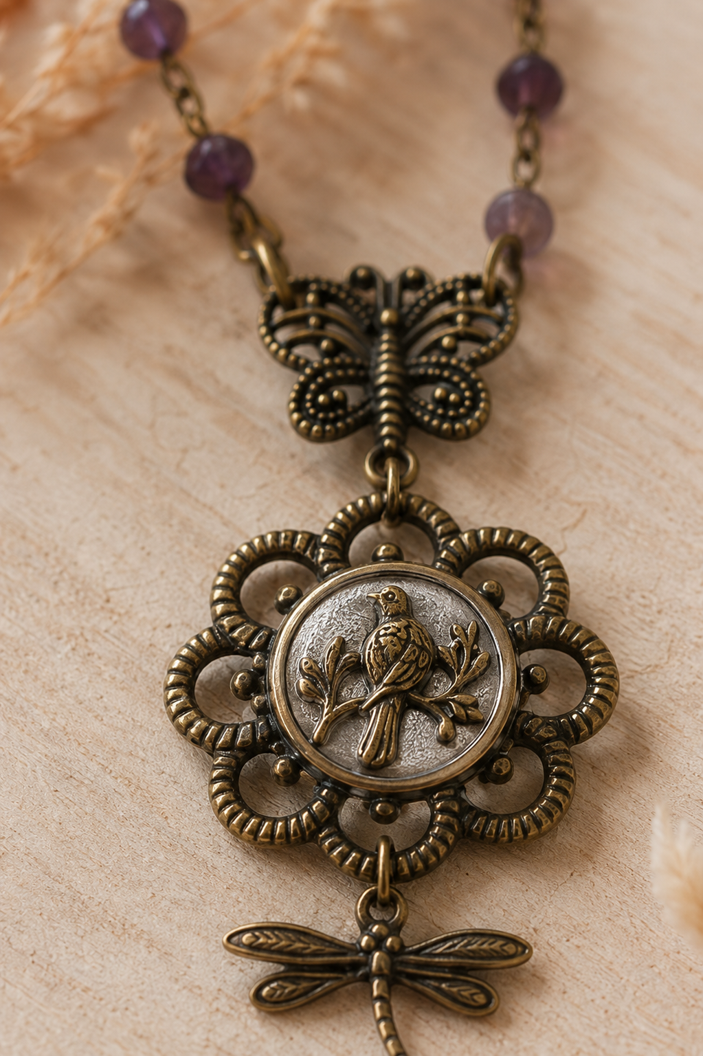 An antique bronze-toned pendant necklace featuring a butterfly, a central oval medallion with a bird motif, and a dangling dragonfly charm, displayed on a light beige textured surface with purple beads at the top.