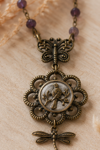 An antique bronze-toned pendant necklace featuring a butterfly, a central oval medallion with a bird motif, and a dangling dragonfly charm, displayed on a light beige textured surface with purple beads at the top.