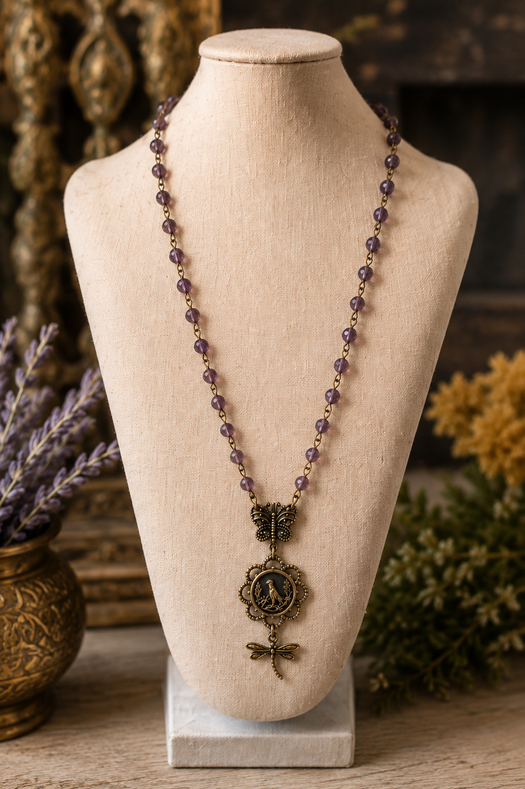 A bohemian-style beaded necklace with purple amethyst beads and an antique bronze pendant featuring a classical bust and dragonfly, displayed on a beige fabric mannequin bust.