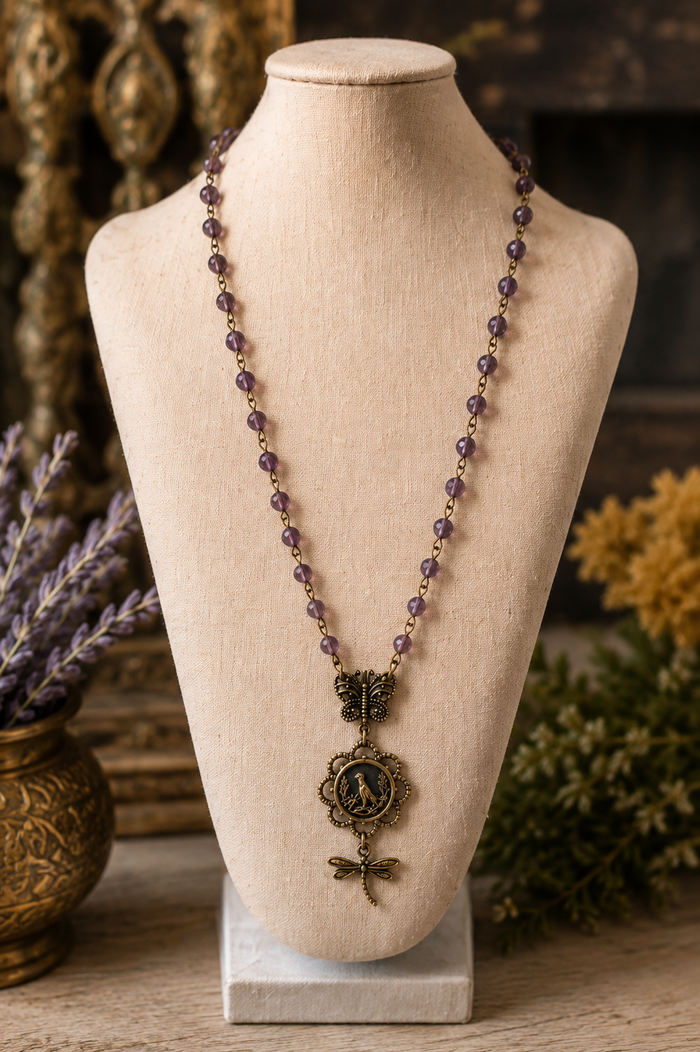 A bohemian-style beaded necklace with purple amethyst beads and an antique bronze pendant featuring a classical bust and dragonfly, displayed on a beige fabric mannequin bust.