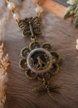 An ornate antique bronze pendant necklace featuring a butterfly and dragonfly filigree design surrounding a central round cabochon with a bird silhouette, displayed on a rustic wooden surface.