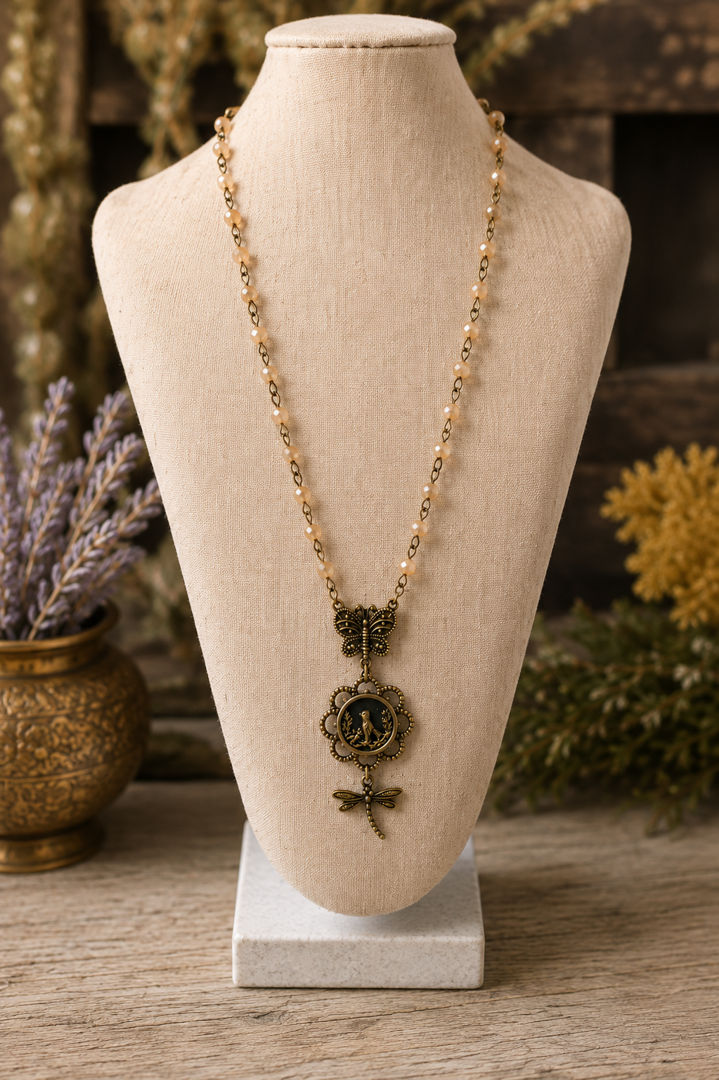 A bohemian-style necklace with a gold-toned beaded chain and a central bronze dragonfly pendant, displayed on a beige fabric mannequin bust.