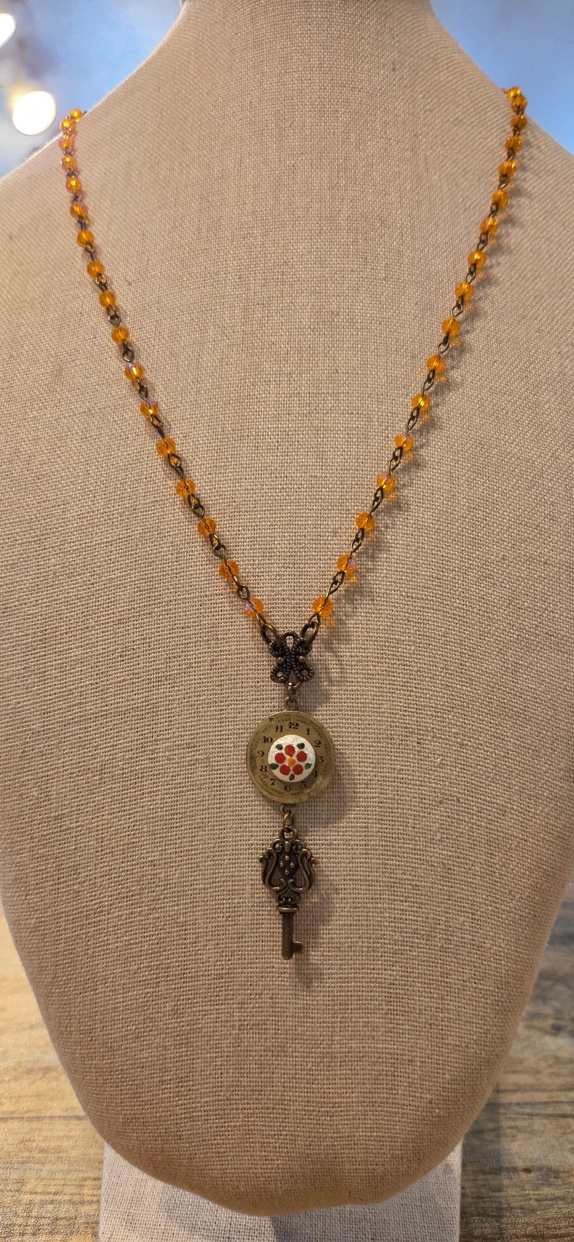 A beaded necklace displayed on a beige fabric mannequin bust. The necklace features a strand of amber-orange and dark metallic beads, a small dark metal key-shaped charm, and a round medallion pendant with a floral design.