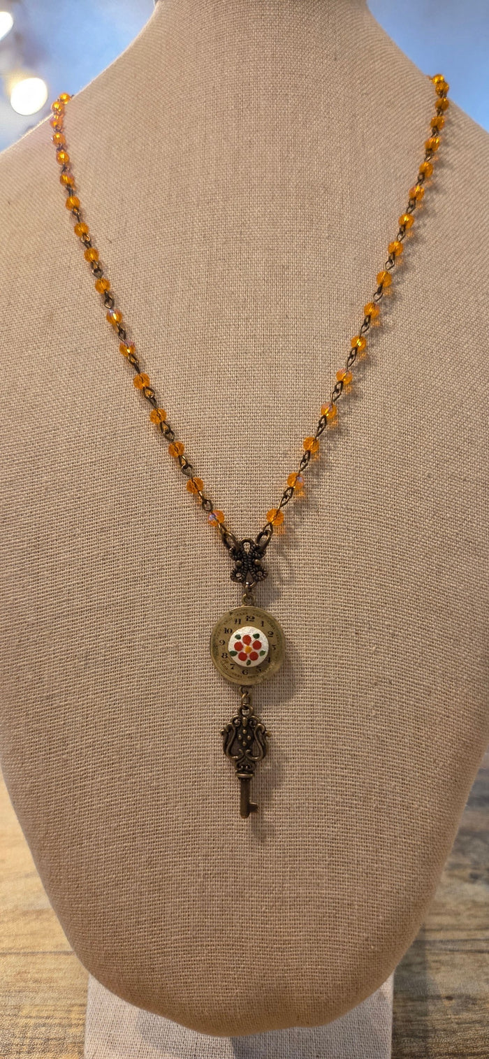 A beaded necklace displayed on a beige fabric mannequin bust. The necklace features a strand of amber-orange and dark metallic beads, a small dark metal key-shaped charm, and a round medallion pendant with a floral design.