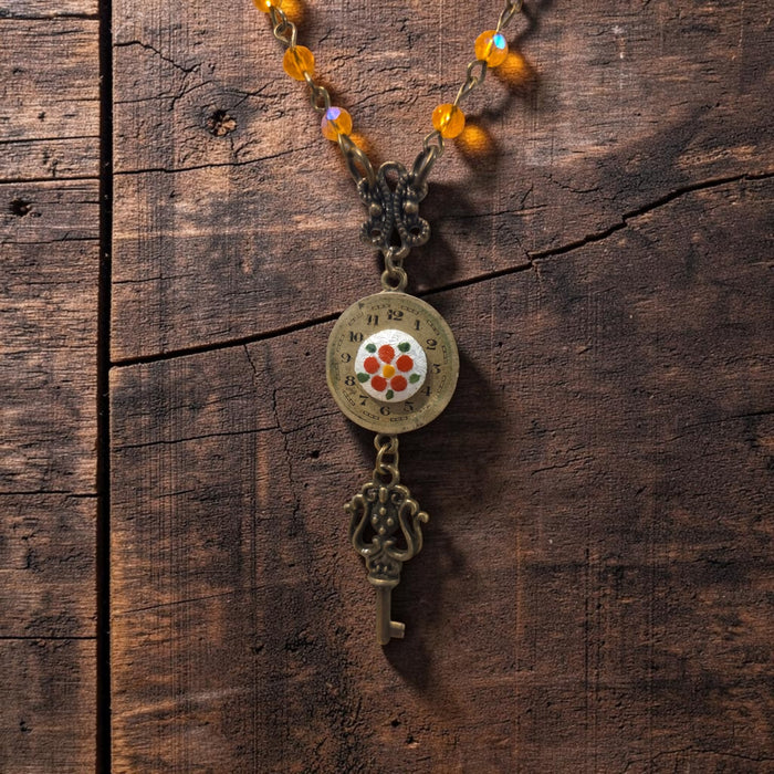A vintage-style key necklace with a round clock face pendant, a floral center, and a hanging key charm, displayed on a rustic dark wood background.