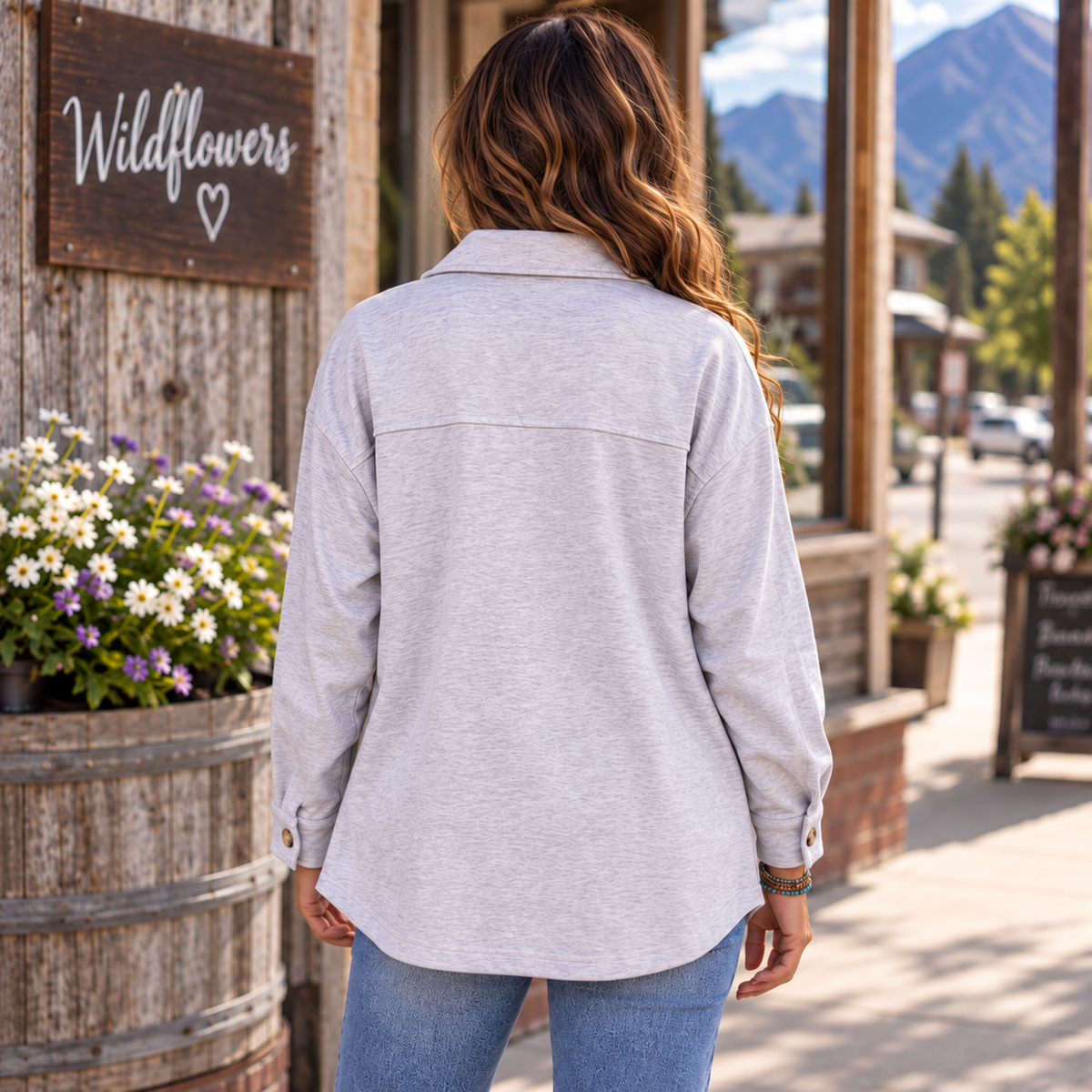 A woman viewed from behind wearing a light heather gray long-sleeve button-up shirt and blue jeans, standing on a sunny outdoor street in front of a rustic wooden building with a sign that reads 'Wildflowers'.