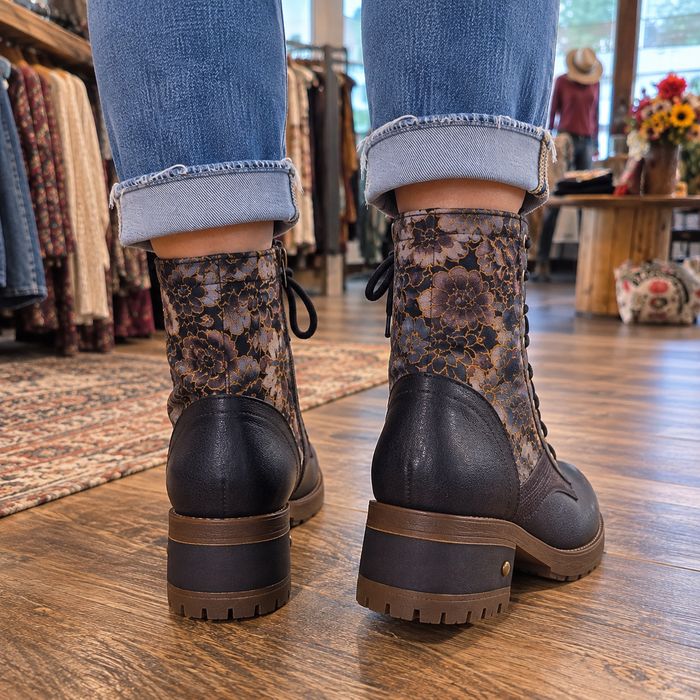 A person wearing cuffed blue jeans and lace-up ankle boots with a floral patterned shaft and dark brown leather heels, standing on a wooden floor inside a boutique.