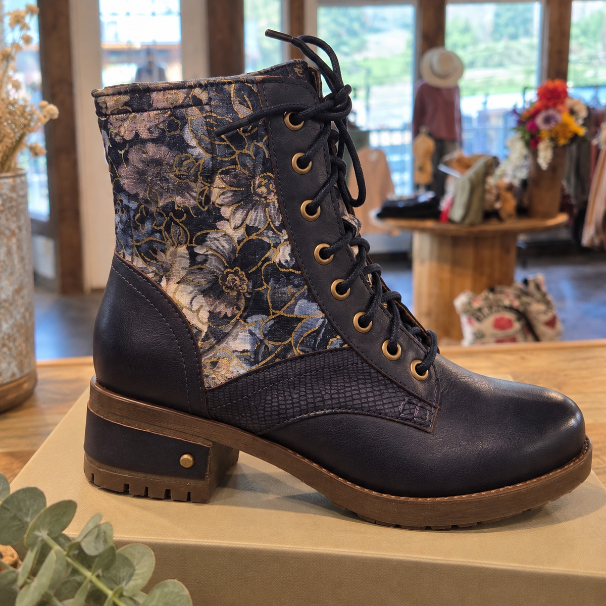 A lace-up ankle boot with a dark navy leather lower section, a textured snakeskin panel, and a floral patterned fabric collar, displayed on a wooden surface in a boutique setting.