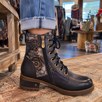 A person wearing a dark brown lace-up ankle boot with floral patterned side panels, a side zipper, and gold-tone eyelets, shown in a retail store setting.