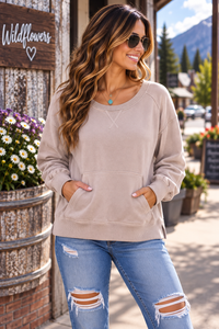 A woman wearing a beige crewneck sweatshirt and distressed blue jeans stands outdoors in front of a rustic wooden sign that reads 'Wildflowers'.