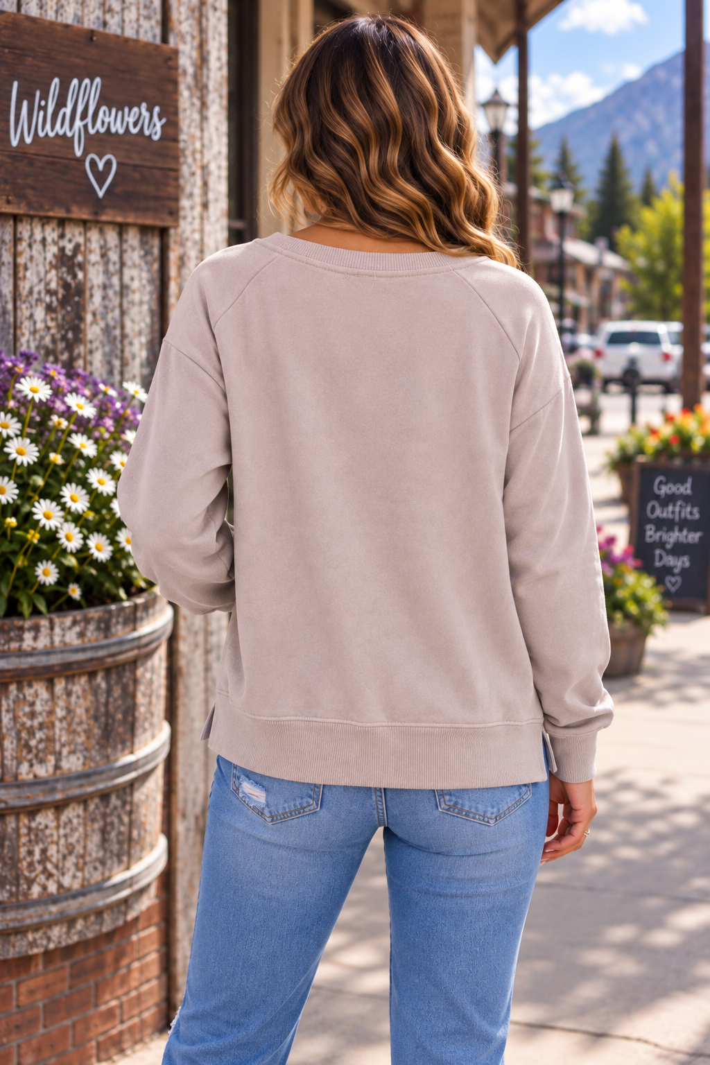 A woman viewed from behind wearing a light taupe crewneck sweatshirt and blue jeans, standing on a sidewalk in front of a rustic wooden building with flowers and mountains in the background.