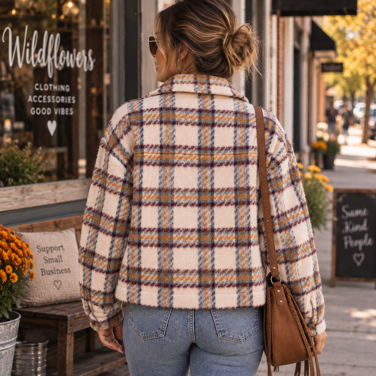 A woman viewed from behind wearing a cream-colored fuzzy plaid jacket with brown, orange, and blue lines, paired with blue jeans and a brown shoulder bag, standing on a sidewalk in front of a storefront.