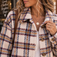 A close-up of a woman wearing a fuzzy plaid coat in cream, mustard yellow, and purple tones, with a white ribbed top underneath and a gold pendant necklace.