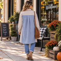 A woman viewed from behind walking on a storefront sidewalk, wearing a long color-block coat with beige and light blue panels, cuffed blue jeans, tan ankle boots, and a brown shoulder bag. The shop window has a sign reading “Wildflowers,” with fall decor including orange flowers, pumpkins, and a bench in the background.