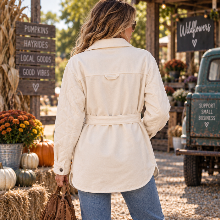 A woman viewed from behind wearing a quilted cream-colored belted jacket in an outdoor autumn farm market setting with pumpkins, flowers, and a vintage truck.
