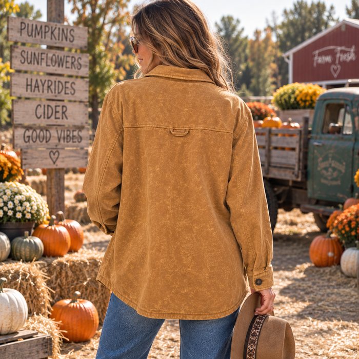 A woman stands outdoors at a fall farm display wearing a mustard brown shirt jacket and blue jeans, holding a brown hat. Pumpkins, hay bales, a rustic sign, and a vintage green truck are visible in the background.