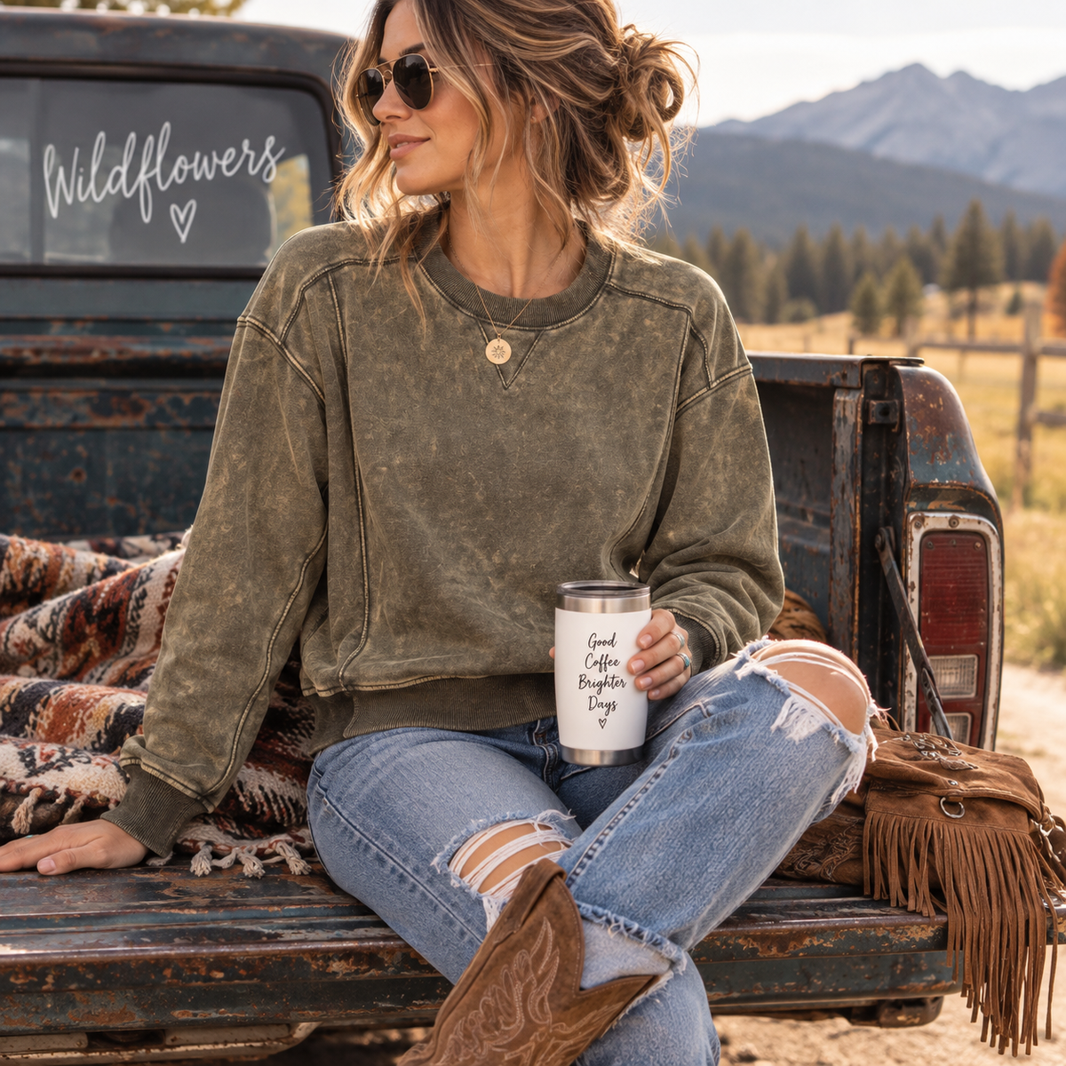 A woman sits on the bed of a rustic pickup truck outdoors, wearing a faded olive green crewneck sweatshirt, distressed blue jeans, and cowboy boots while holding a white travel tumbler with black text.