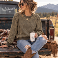 A woman sits on the bed of a rustic pickup truck outdoors, wearing a faded olive green crewneck sweatshirt, distressed blue jeans, and cowboy boots while holding a white travel tumbler with black text.