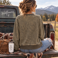 A woman sits in the bed of a pickup truck facing mountains, wearing a washed olive green crewneck sweatshirt and blue jeans. A white insulated tumbler with text sits on a patterned blanket next to her.