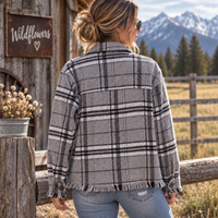 A woman stands outdoors in a rustic mountain setting wearing a gray, black, and white plaid flannel shirt jacket with fringed edges and light blue jeans.