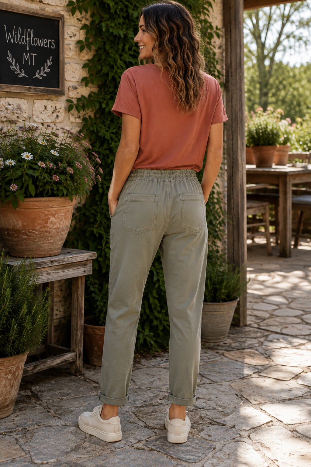 A woman stands on a stone patio wearing light sage green drawstring pants, a terracotta t-shirt, and white sneakers, photographed from behind in an outdoor rustic setting.
