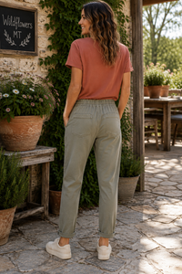 A woman stands on a stone patio wearing light sage green drawstring pants, a terracotta t-shirt, and white sneakers, photographed from behind in an outdoor rustic setting.