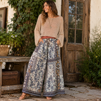 A woman stands outdoors in front of a rustic wooden door and stone wall, wearing a beige knit cropped sweater and high-waisted, wide-leg boho pants with a blue, beige, and brown floral pattern.