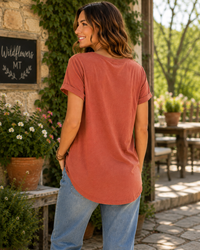 A woman wearing a dusty terracotta short-sleeve top and light blue jeans, photographed from behind in an outdoor patio setting with stone walls, potted plants, and greenery.